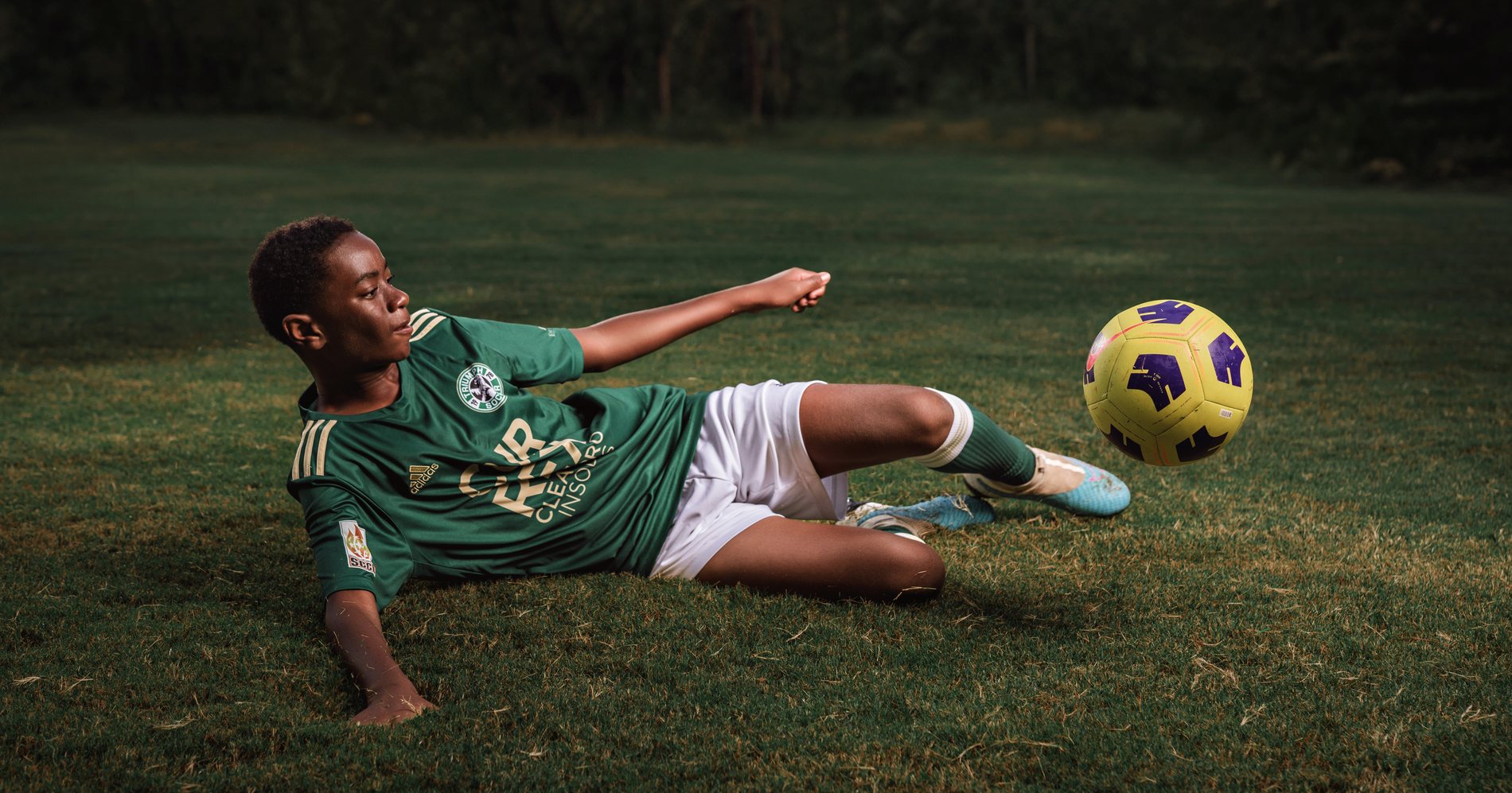 Soccer player in a green kit sliding low on the grass to strike a yellow ball