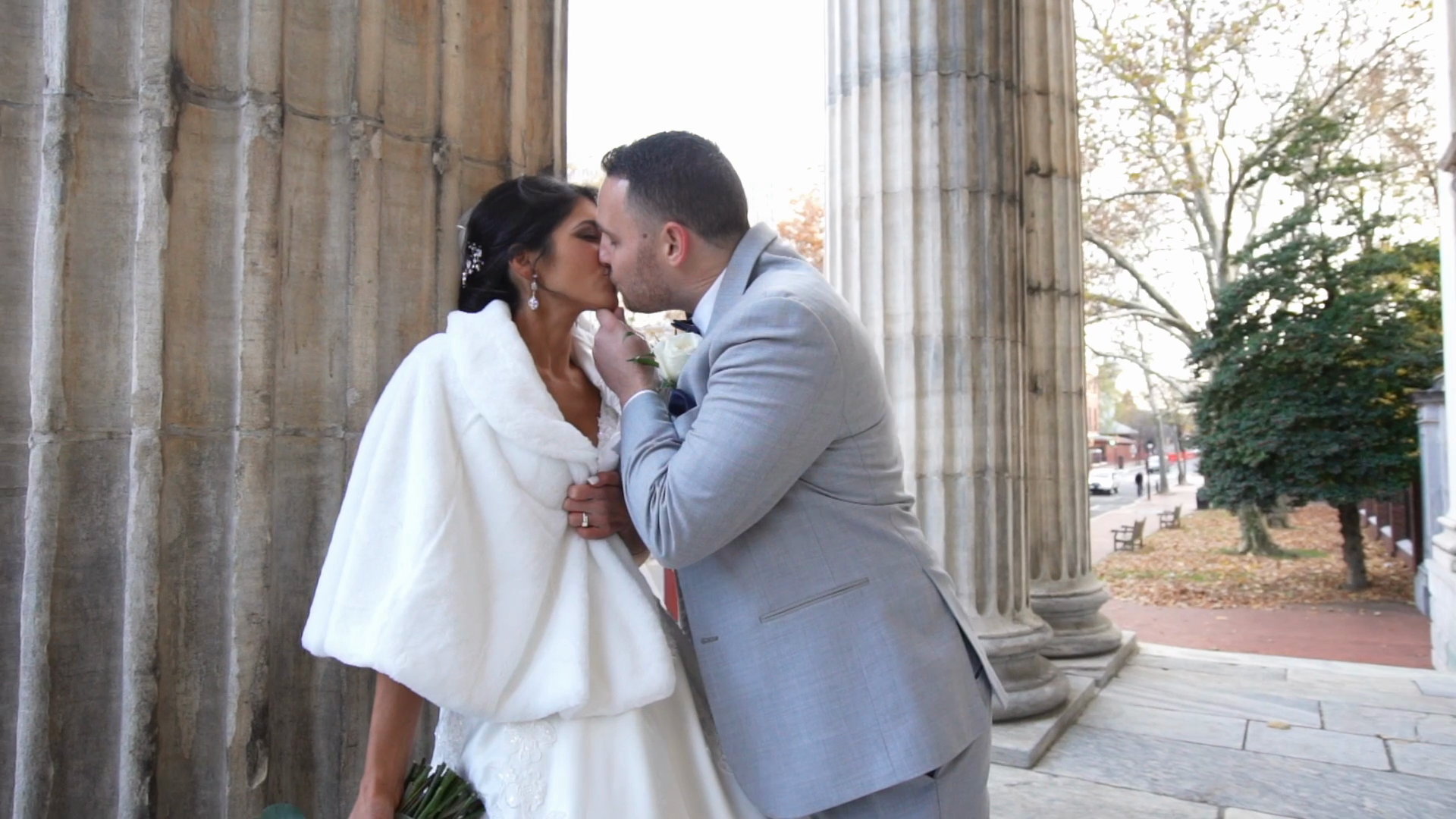 Alyssa and Will kissing beside a fluted stone column, Alyssa in a white fur wrap